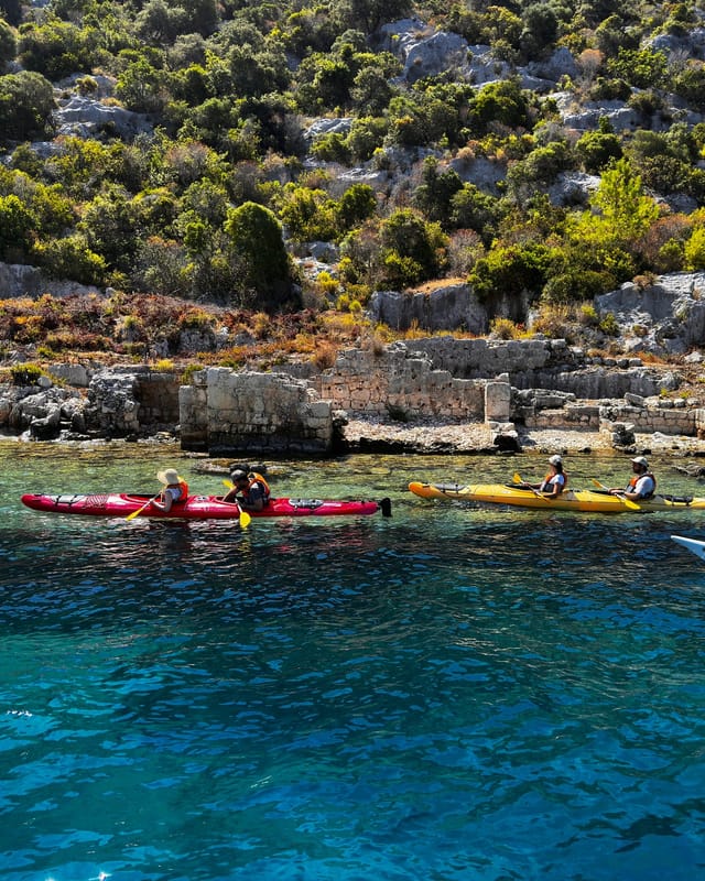 The Sunken City shoreline: ancient walls running down into the water, red and yellow sea kayaks passing in front of them