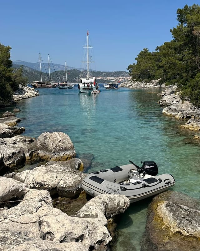 Çamlık Cove: clear shallow water between the rocks, an inflatable tender pulled up on the stone, gulets at anchor beyond