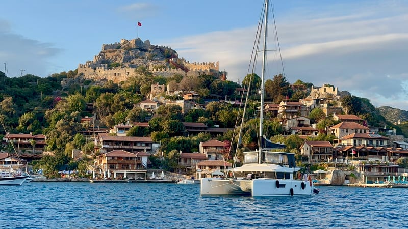 Kaleköy from the water: Simena Castle on the hilltop with a Turkish flag on the wall, stone houses down the slope, a white sailing catamaran at anchor in front